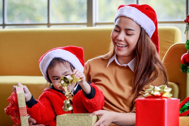 Happy Asian family daughter girl wears sweater red and white Santa Claus hat sitting with mom surprising exciting when open present gift box celebrate Xmas eve near Christmas pine tree in living room.