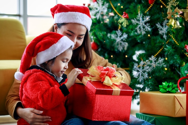 幸せなアジアの家族の娘の女の子は、自宅のリビングルームのクリスマス松の木の近くでクリスマスイブを祝うお母さんの箱を開けるオープンプレゼントギフトボックスと一緒に座っている赤と白のセーターのサンタクロースの帽子をかぶっています。