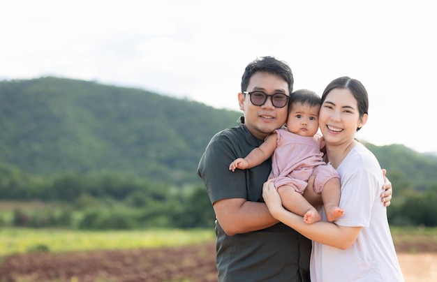 自然の背景に幸せなアジアの家族の父母子娘。両親は晴れた日に屋外に立っている腕の中で小さな赤ちゃんを抱きしめます。