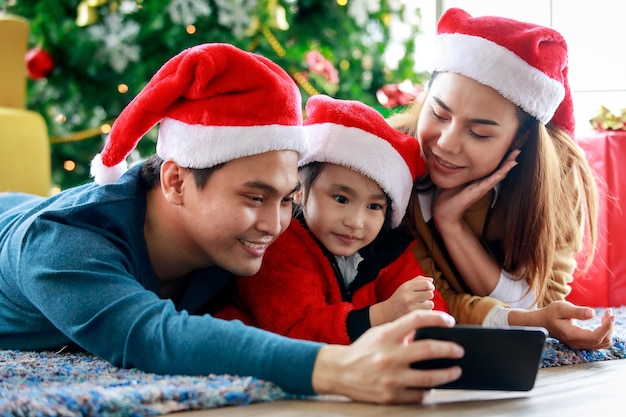 幸せなアジアの家族の父の母と娘は、一緒にクリスマスイブを祝う自分撮り写真を撮るスマートフォンのカメラを使用してカーペットの床に横たわる赤と白のサンタクロースの帽子とセーターを着ています。