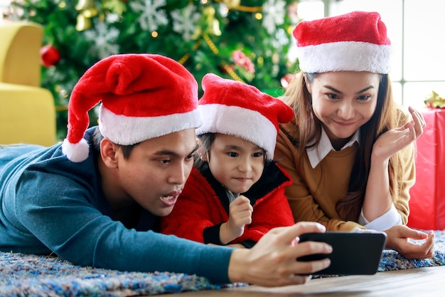 幸せなアジアの家族の父の母と娘は、一緒にクリスマスイブを祝う自分撮り写真を撮るスマートフォンのカメラを使用してカーペットの床に横たわる赤と白のサンタクロースの帽子とセーターを着ています。