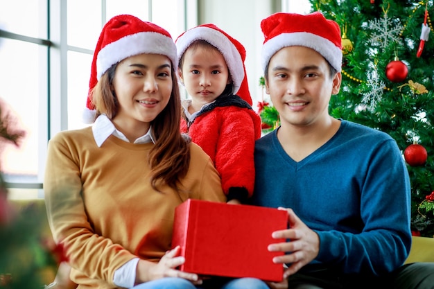 Happy Asian family father mother and daughter wears sweater with red and white Santa Claus hat smiling laughing holding present gift box prepare to unbox open it together celebrating Christmas eve.