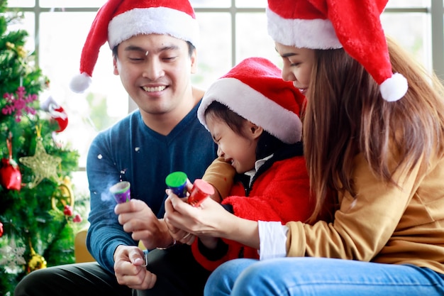 Happy asian family father mother sitting on sofa helping afraid daughter wears sweater with red and white santa claus hat pulling exploding party popper confetti together celebrating christmas eve