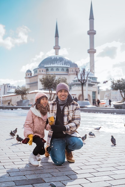 Photo happy asian family feeding pigeons in city sqare with mosque in the background