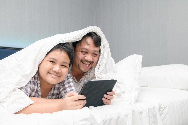 Happy asian family having fun with device at home, father and\
his son using digital tablet looking at screen lying on bed\
together,