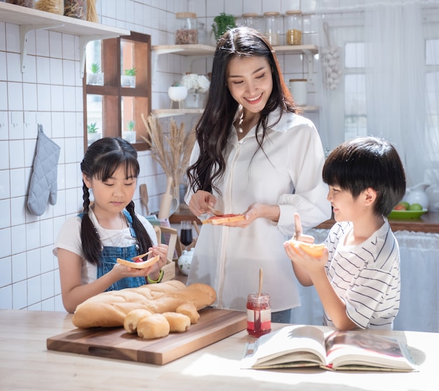 Happy asian family in the kitchen. mother and son and daughter spread strawberry yam on bread, leisure activities at home.