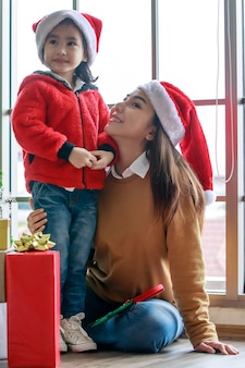 Felice famiglia asiatica mamma e figlia in cappello di babbo natale rosso e bianco e maglione sorridente che giocano insieme vicino all'albero di pino di natale con elementi decorativi glitterati e scatole regalo regalo nella sera di natale.