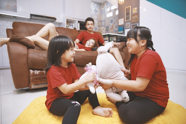 Happy asian family spend free time together in living room at home Couple sitting and laying on sofa looks at two little daughter playing doll on the carpet