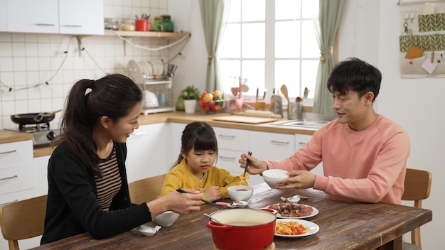 3명의 행복한 아시아 가족이 집에 있는 식당에서 식사를 합니다. 엄마가 앉아서 아기 딸에게 무엇을 먹고 싶은지 묻는 동안 아빠는 접시를 식탁에 놓는 것을 돕습니다.