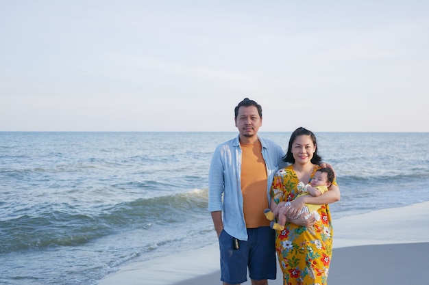 Happy Asian family vacation, Mom and Dad hold a cute baby at the beach in summer, look at camera, family sea trip