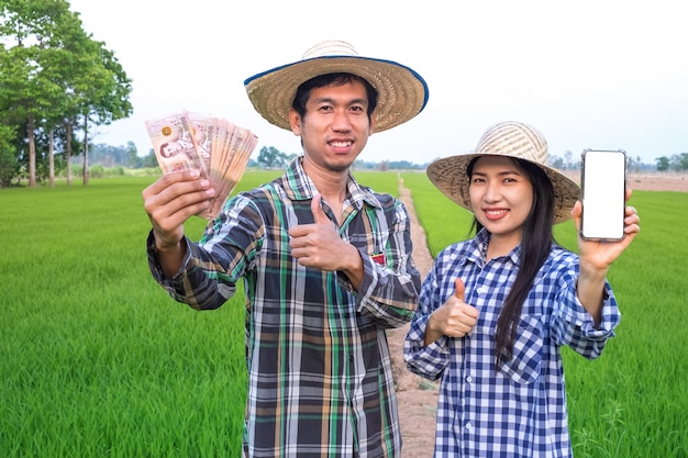 Happy asian farmer couple holding Thai banknote and smartphone with blank screen standing at rice farm