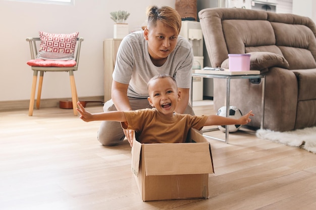 幸せなアジア人の父親が段ボール箱を運転し、興奮した幼い息子が中で子供と遊んでいる父親