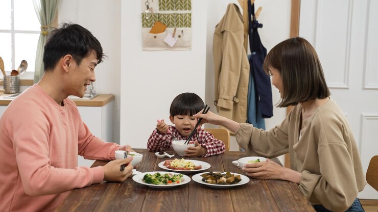 Photo 幸せなアジア人の父、母、就学前の男の子が家で一緒に昼食をとっている。愛情のあるカップルはお互いに食べ物を与え、息子のボウルを与えます