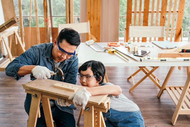 Happy Asian father and son work as a woodworker and carpenter Father teaching his son to hammer nails on a wooden plank carefully together carpentry working at a home workshop studio