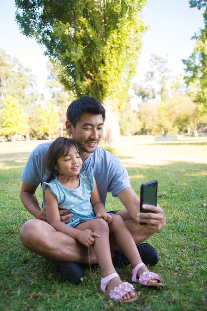 Happy Asian father taking selfie with daughter in public park. Young man resting on green field with little girl on knees both sitting looking at camera.  Leisure with child, new technologies concept