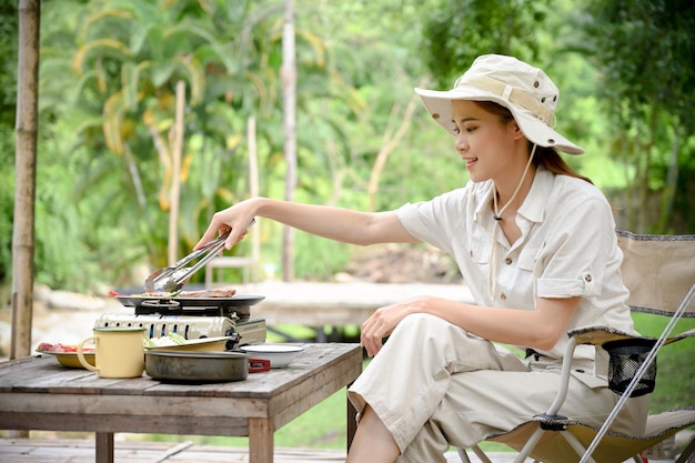 Happy Asian female camping at the campground preparing grilling steak and barbecue