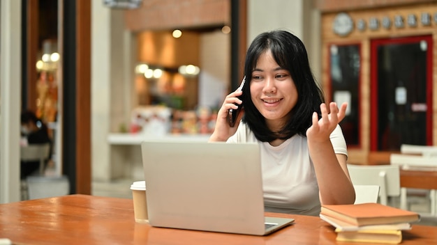A happy Asian female freelancer is on the phone with her client while remote working at a cafe