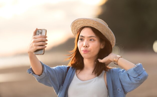 写真 夕日の美しいビーチでの旅行中にスマートフォンで幸せなアジアの女子学生の自撮り写真