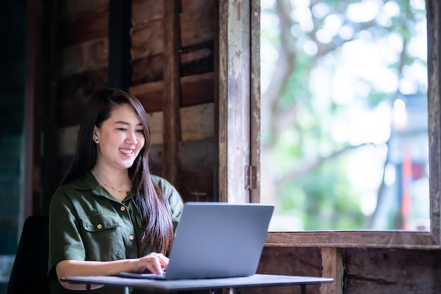 갈색 녹색 드레스를 입고 세련된 힙스터 캐주얼을 입은 아시아 프리랜서 비즈니스 여성이 목조 주택 배경의 창문 옆에 커피 컵이 있는 노트북 컴퓨터와 함께 일하는 것에 만족합니다.