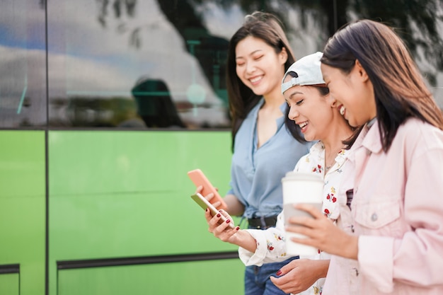 バスの駅でスマートフォンを使用して幸せなアジアの友人。放課後の電話アプリを楽しんでいる若い学生の人々