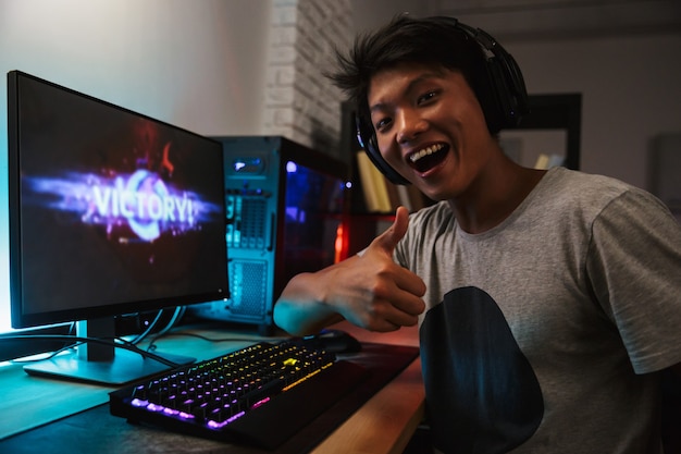 Happy asian gamer guy 16-18 rejoicing victory while playing video games on computer in dark room, wearing headphones and using backlit colorful keyboard