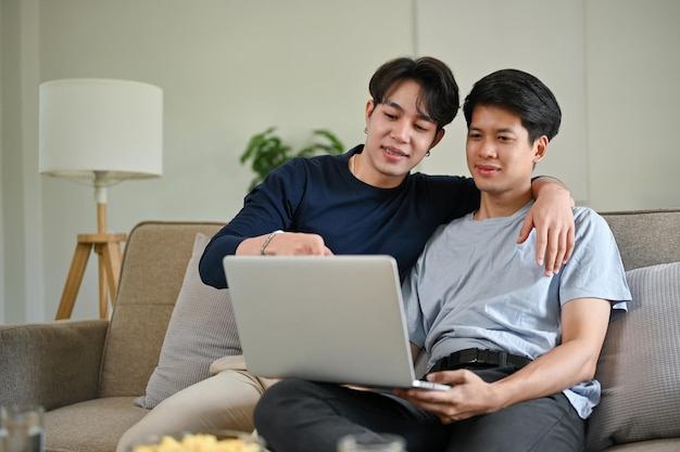 a happy asian gay man enjoys watching a movie through a laptop with his boyfriend on a couch