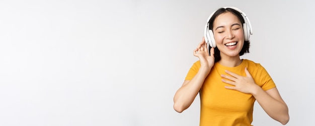 ヘッドフォンで音楽を聴いて踊り、黄色のTシャツに立って笑顔で踊る幸せなアジアの女の子...