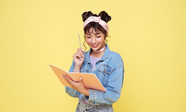 Happy Asian girl holding pencil and notebook over yellow background. back to school concept.