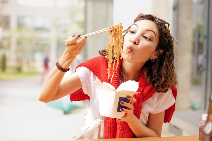 Photo 幸せなアジアの女の子が中国製のチョップスティックを握り,外食用紙箱からウォックヌードルを食べています. ストリートとファストフードのコンセプト