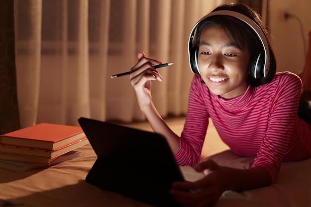 Happy asian girl is studying video conference online with\
teacher on the tablet in the bedroom at night asia child watching\
and education distance on the computer concept learning at\
homexd