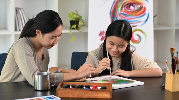 Happy asian girl painting picture with teacher at art class.