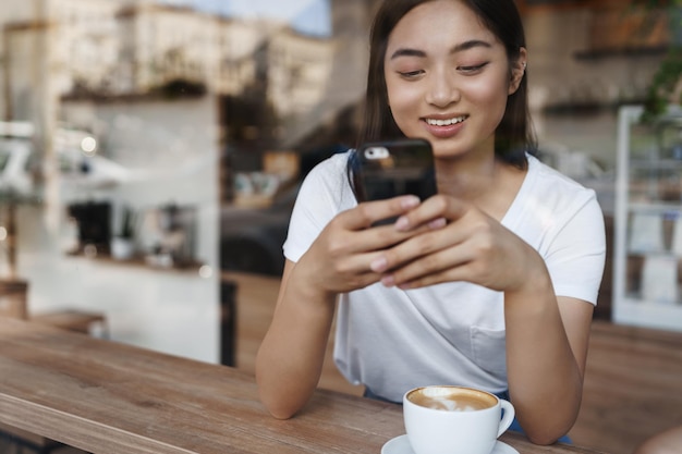 携帯電話でおしゃべりとコーヒーを飲みながらカフェで窓際に座っている幸せなアジアの女の子スマートフォンを使用してレストランでカプチーノを持っている若い女性