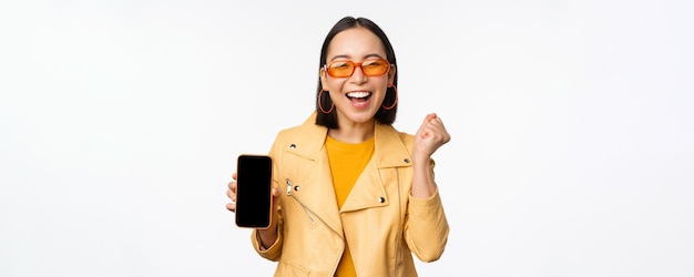 Photo happy asian girl in sunglasses showing mobile phone screen smartphone interface laughing and smiling