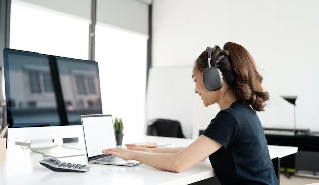 Счастливая азиатская девушка с наушниками, использующая ноутбук, общается онлайн и проводит видеоконференции в домашнем офисе.