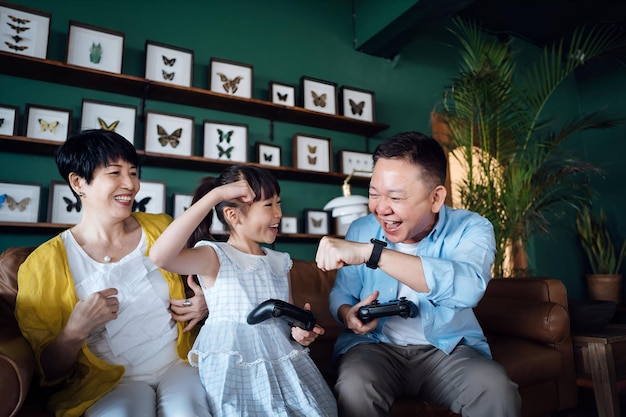 Happy Asian grandparents and granddaughter having fun playing video games together sitting on the sofa in the living room at home Happy retirement concept