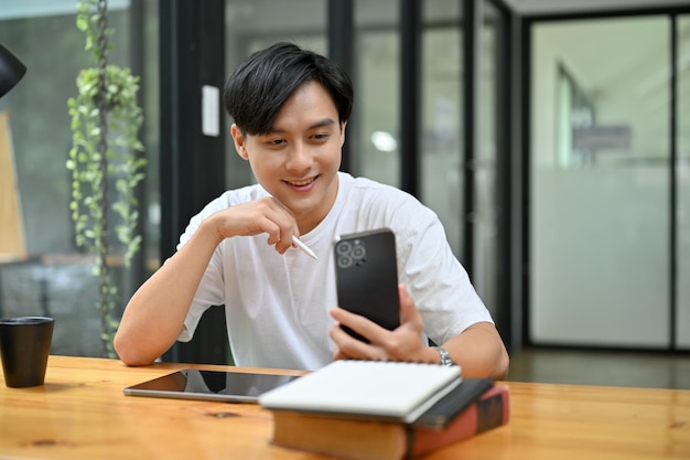 Happy asian guy sits at his working desk using his smartphone to chat with his friends