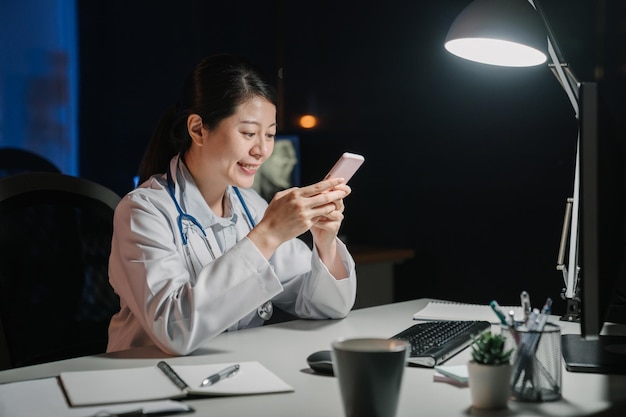 Happy asian japanese female medical doctor sitting at her table and browsing online website on mobile phone at late night. beautiful smiling woman nurse staff in dark clinic office using cellphone.