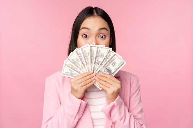 Happy asian lady in suit holding money dollars with pleased face expression standing over pink background Copy space