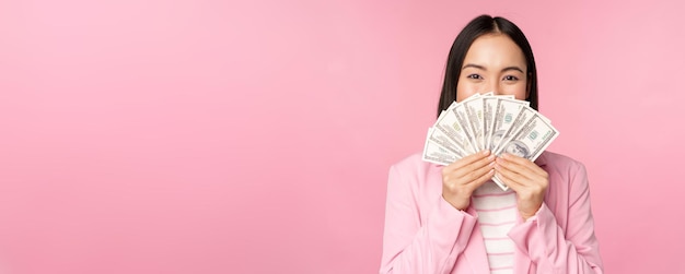 ピンクの背景の上に立っている満足のいく表情でお金のドルを保持しているスーツの幸せなアジアの女性コピースペース