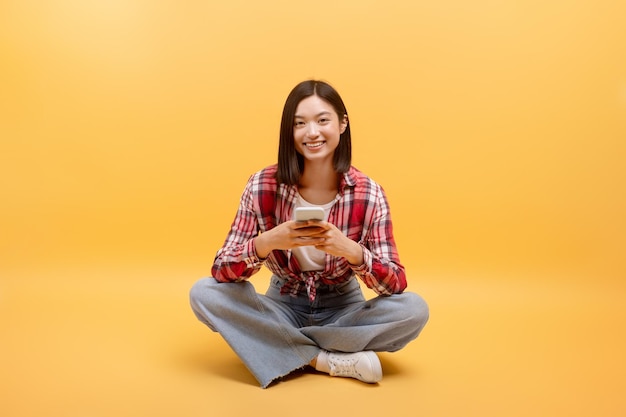 Happy asian lady using cellphone texting or browsing internet sitting on floor over yellow