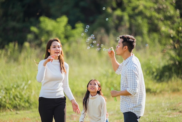 Happy Asian lifestyle family mother, father and little cute girl child having fun together and enjoying outdoor play blowing soap bubbles in the garden park on a sunny day, summertime