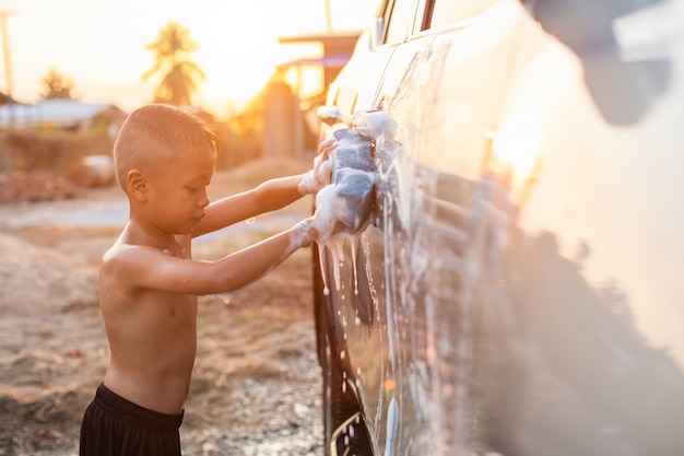 Happy asian little boy with white soap and using blue sponge to washing the car at outdoor in sunset time