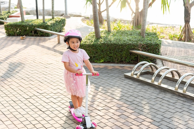 Happy Asian little kid girl wear safe helmet playing pink kick board on road in park outdoors on summer day