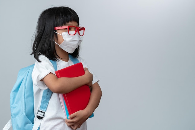 Happy asian little preschool girl wearing red glasses holding books and red backpack