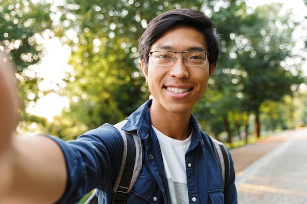 Photo happy asian male student in eyeglasses making selfie and looking at th camera outdoors