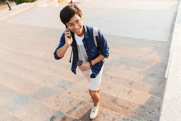 Happy asian male student in eyeglasses talking by smartphone while looking away and walking on stairs outdoors