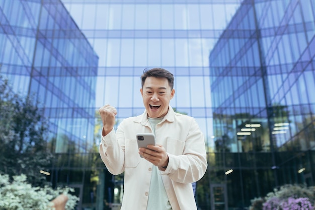 Happy asian man celebrating victory and success holding phone smiling and happy programmer outside