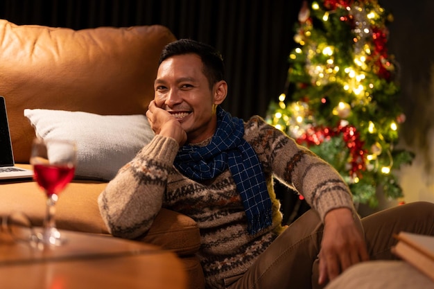 Happy asian man in comfy winter clothes sitting beside a sofa in his living room on christmas night