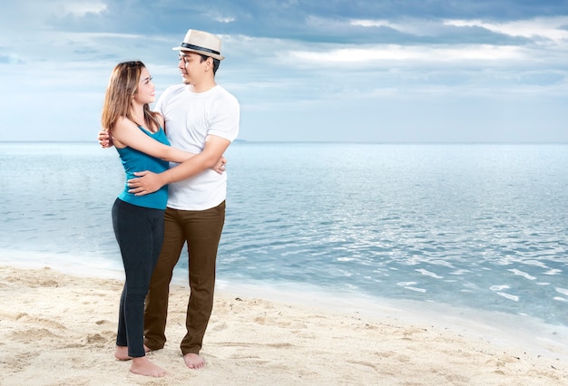 Happy asian man in hat hugging his girlfriend on the beach