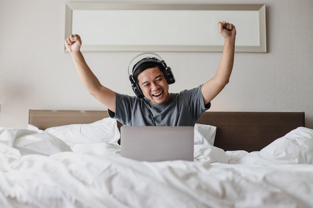 헤드폰을 끼고 침대에서 승리의 제스처를 표현하는 흥분된 표정으로 노트북을 들여다보는 행복한 아시아 남자